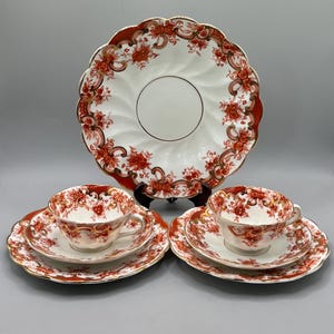 May include: A porcelain tea set consisting of one large serving plate, two teacups, and two saucers. Each piece features a white base with a scalloped edge, decorated with a vibrant orange-red floral pattern and gold trim accents.