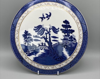 May include: A blue and white porcelain plate with a gold rim and a willow pattern design. The plate features a willow tree, a bridge, a pagoda, and two birds in flight.