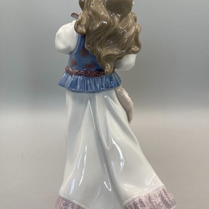 Puede incluir: Figurita de porcelana de una ni&ntilde;a con un vestido azul y blanco con estampado floral. Tiene el pelo largo y casta&ntilde;o y lleva una corona de flores. La figurita est&aacute; de pie sobre un fondo blanco.