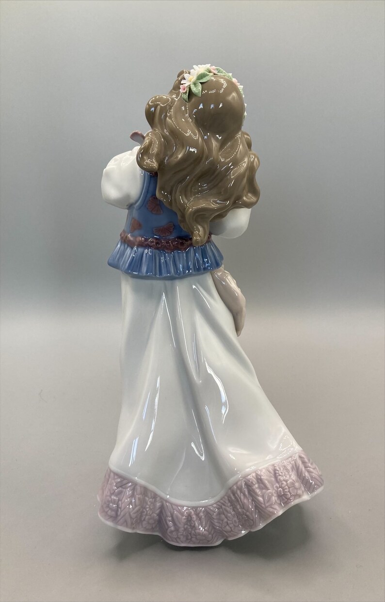 Puede incluir: Figurita de porcelana de una ni&ntilde;a con un vestido azul y blanco con estampado floral. Tiene el pelo largo y casta&ntilde;o y lleva una corona de flores. La figurita est&aacute; de pie sobre un fondo blanco.