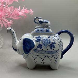 May include: A white ceramic teapot shaped like an elephant with blue painted floral and geometric patterns. The lid features a small sculpted elephant figurine as a handle. The teapot has a curved spout, a blue handle, and four sturdy legs.