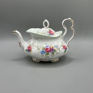 Könnte beinhalten: Eine weiße Porzellan-Teekanne mit einem bunten Blumenmuster aus rosa Rosen, blauen Kornblumen und violetten Blüten. Die Teekanne hat einen gewellten Rand, einen geschwungenen Henkel, einen Ausguss und einen Deckel mit ringförmigem Knauf.