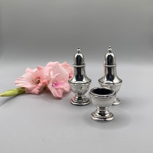 Könnte beinhalten: Ein Set aus drei silbernen Salz- und Pfefferstreuern. Die Streuer sind rund und haben ein Perlenmuster um den oberen und unteren Rand. Die Streuer stehen auf einem weißen Hintergrund.