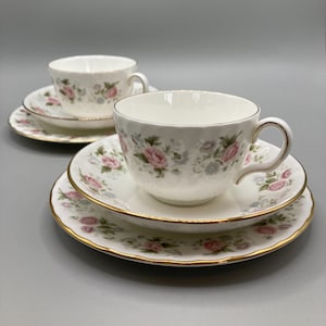 May include: Two white teacups and saucers with a floral pattern in shades of pink, blue, and green. The cups and saucers have a gold trim.