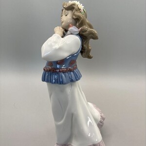 Puede incluir: Una figurita de porcelana de una ni&ntilde;a que lleva un vestido blanco con un chaleco azul y rojo, una corona de flores y sostiene un p&aacute;jaro peque&ntilde;o.