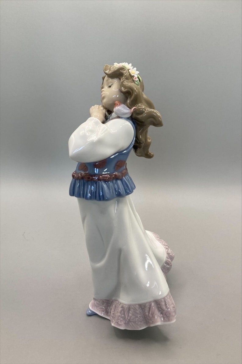Puede incluir: Una figurita de porcelana de una ni&ntilde;a que lleva un vestido blanco con un chaleco azul y rojo, una corona de flores y sostiene un p&aacute;jaro peque&ntilde;o.