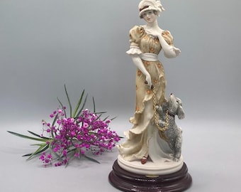 May include: A porcelain figurine of a woman in a floral dress holding a leash attached to a small poodle. The figurine is standing on a wooden base.