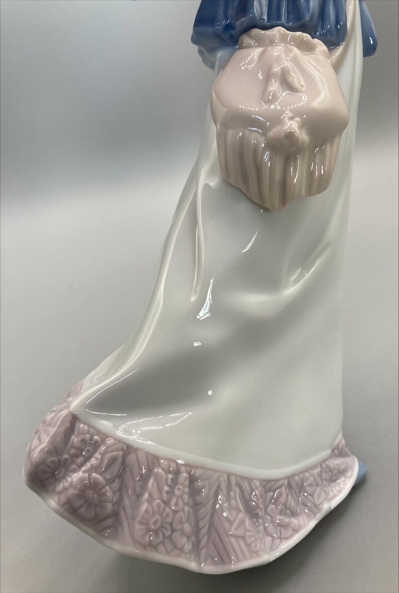 Puede incluir: Una figurita de porcelana de una mujer con un vestido azul y blanco con un bolso rosa. El vestido tiene un ribete floral rosa.