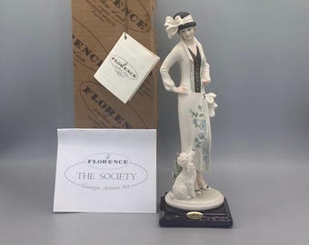 Könnte beinhalten: Eine Porzellanfigur einer Frau in einem weißen Kleid und Hut, die einen weißen Hund hält. Die Figur steht auf einem schwarzen Sockel und trägt ein Etikett mit der Aufschrift "Florence The Society Giuseppe Armani Art".