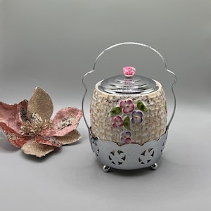 Pode incluir: Uma açucareira vintage com tampa e alça prateadas, e uma base decorativa prateada. A tigela de cerâmica é bege com um padrão texturizado e detalhes florais em rosa, roxo e verde. Um adorno floral rosa está à esquerda.