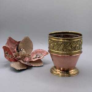 Peut inclure: Un récipient décoratif en cuivre et laiton avec un motif floral. Le récipient a un bord en laiton avec un motif floral en relief et un corps en cuivre. Une grande fleur rose et pailletée est à côté du récipient.