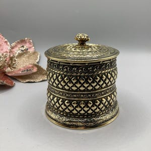 Op de afbeelding: Oud metalen pot met deksel in antieke stijl. De pot is goudkleurig met ingewikkelde reliëfpatronen, waaronder een rasterontwerp rond de body en bloemendetails op het deksel en de basis. Het deksel heeft een kleine, decoratieve knop.