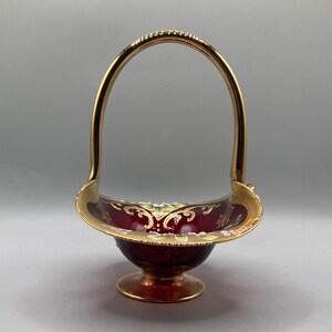 Puede incluir: Una cesta de vidrio roja con un asa dorada y detalles florales pintados. La cesta tiene una base redonda y está decorada con ribetes dorados.