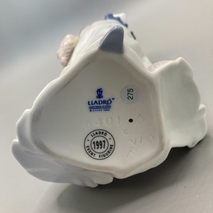 Puede incluir: Figurita de porcelana blanca con detalles azules. La base de la figurita tiene una etiqueta azul con el texto "LLADRO HAND MADE IN SPAIN O DAISA 1999" y un c&iacute;rculo azul con el texto "1997 EVENT FIGURINE". Tambi&eacute;n hay un c&iacute;rculo azul con el n&uacute;mero "275" y un c&iacute;rculo azul con el n&uacute;mero "401".