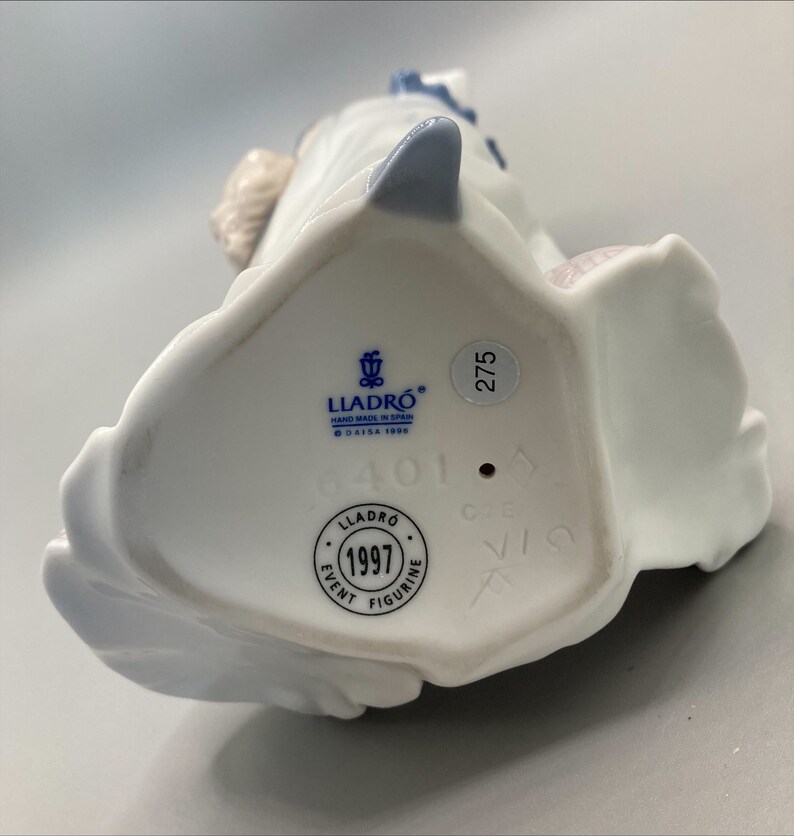 Puede incluir: Figurita de porcelana blanca con detalles azules. La base de la figurita tiene una etiqueta azul con el texto "LLADRO HAND MADE IN SPAIN O DAISA 1999" y un c&iacute;rculo azul con el texto "1997 EVENT FIGURINE". Tambi&eacute;n hay un c&iacute;rculo azul con el n&uacute;mero "275" y un c&iacute;rculo azul con el n&uacute;mero "401".