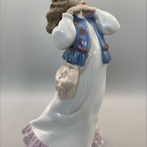 Puede incluir: Una figurita de porcelana de una ni&ntilde;a con cabello casta&ntilde;o que lleva un vestido blanco con un chaleco azul y una diadema floral. Ella se est&aacute; sosteniendo las manos en la barbilla y tiene una peque&ntilde;a bolsa colgando de su cintura.