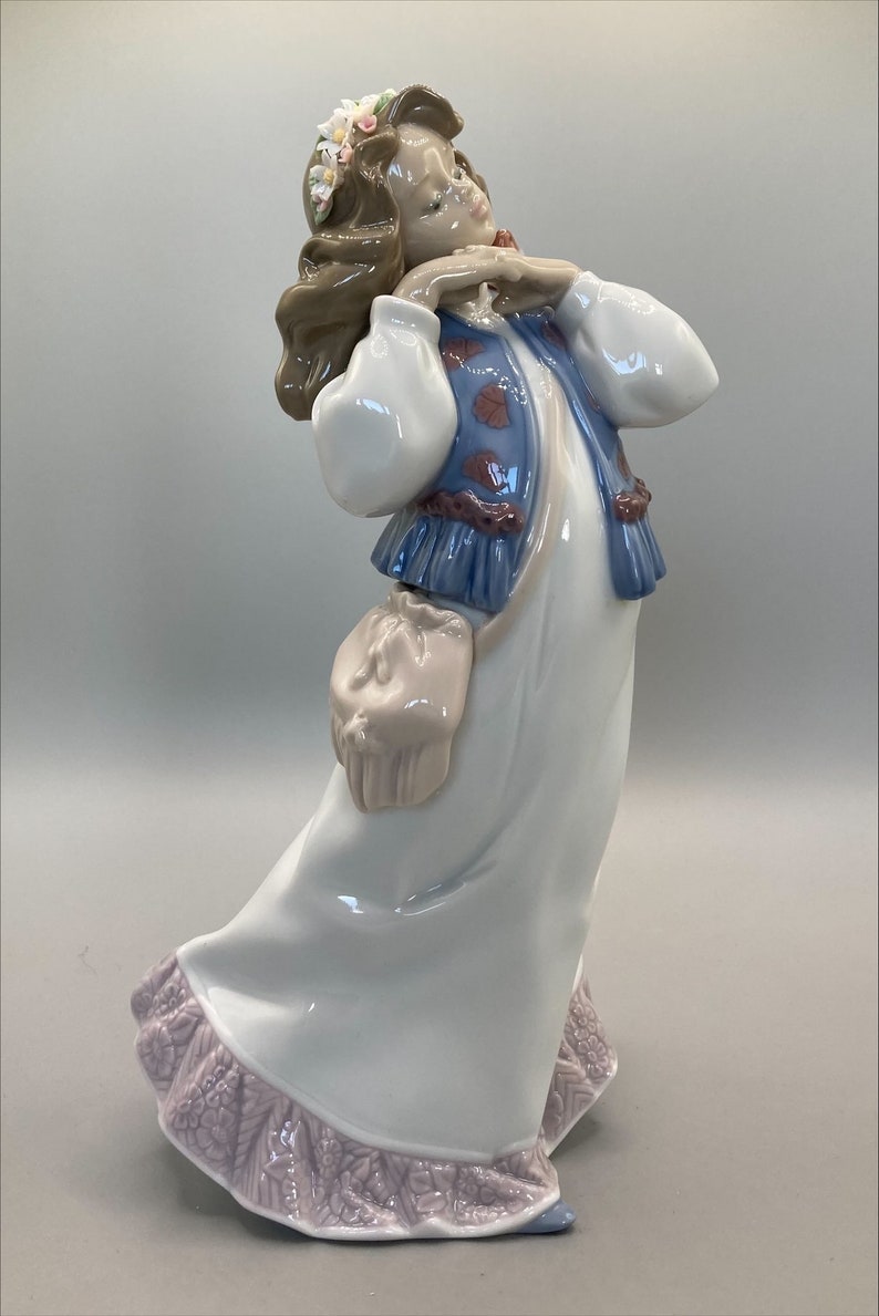 Puede incluir: Una figurita de porcelana de una ni&ntilde;a con cabello casta&ntilde;o que lleva un vestido blanco con un chaleco azul y una diadema floral. Ella se est&aacute; sosteniendo las manos en la barbilla y tiene una peque&ntilde;a bolsa colgando de su cintura.