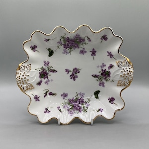 May include: White porcelain serving dish with a scalloped edge and gold trim. The dish is decorated with clusters of purple violets and green leaves. The dish has a decorative gold design on each end.