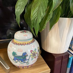 Peut inclure: Un pot à gingembre en céramique blanche avec un couvercle, orné d'un motif coloré représentant une créature mythique bleue, une roue jaune et rose et d'autres éléments décoratifs. Le pot est rond avec un couvercle.