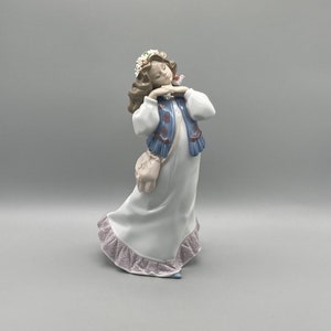 Puede incluir: Una figurita de porcelana de una ni&ntilde;a que lleva un vestido blanco con un chaleco azul y una corona de flores. Est&aacute; sosteniendo un peque&ntilde;o p&aacute;jaro en su mano y tiene una bolsa rosa colgada del hombro.