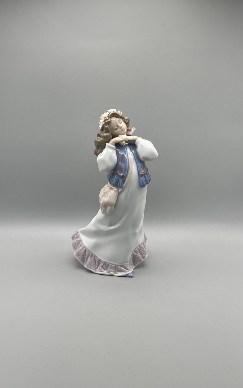 Puede incluir: Una figurita de porcelana de una ni&ntilde;a que lleva un vestido blanco con un chaleco azul y una corona de flores. Est&aacute; sosteniendo un peque&ntilde;o p&aacute;jaro en su mano y tiene una bolsa rosa colgada del hombro.
