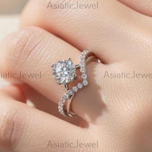 May include: A diamond engagement ring with a round-cut center stone and a curved band set with smaller diamonds. The ring is on a hand, with a soft, blurred background. The ring is a silver color.