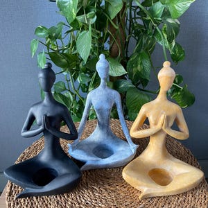 Op de afbeelding: Drie yogapose-beeldjes in zwart, blauw en goud. Elk beeldje zit met de handen in gebedshouding. De beeldjes staan op een geweven mat, met groene planten op de achtergrond.
