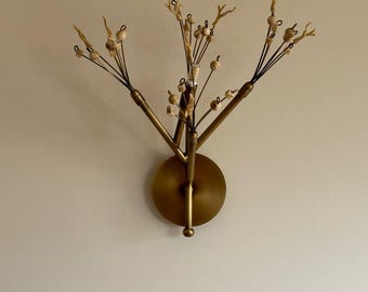 May include: A brass wall sconce with a tree-like design. The sconce features a circular base and multiple branches adorned with small, light-colored beads. The branches extend outward, creating a decorative and artistic lighting fixture.