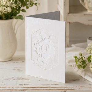 May include: A white folded card featuring a die-cut rectangular frame with scalloped edges on the front cover. The centre of the card displays a light pink monogram with the letters OP, and a small white pearl accent is positioned at the bottom.