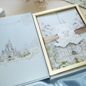 May include: A light blue wedding invitation box with a gold trim. The box features a watercolor illustration of Cinderella's castle and a floral design. The inside of the box has a white card with the couple's initials and wedding date.