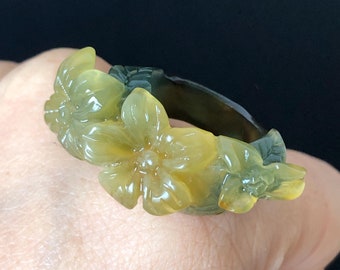 以下が含まれることがあります： 花柄の彫刻が施された翡翠の指輪。指輪は緑色の翡翠でできており、表面に黄色の花が彫刻されています。花は指輪の周りに円形に配置されています。