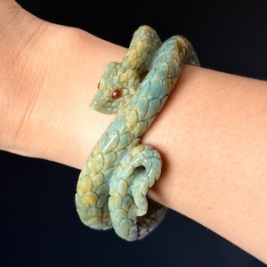 May include: A coiled snake-shaped bracelet featuring a textured, scale-like surface in shades of muted green and brown. The serpent design includes a detailed head with small, dark red inlaid eyes, wrapping around the wrist in a continuous loop.