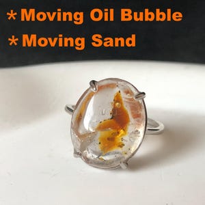 Op de afbeelding: Een heldere, ovale ring met een zilveren band. De ring heeft een oranje en gele insluitsel. De tekst "Moving Oil Bubble" en "Moving Sand" is zichtbaar op de achtergrond.