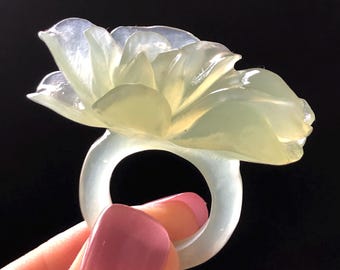 以下が含まれることがあります： 透明感のある、淡い緑色のフラワーリング。リングは、複数の花びらを持つ、詳細なオープンフラワーデザインが特徴です。リングは黒い背景に対して保持され、花の繊細なディテールを示しています。
