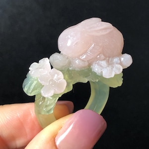 May include: A carved ring featuring a pink bunny atop a floral design. The ring has a light green band with white flowers and leaves. The bunny is a soft pink color, and the flowers are white.