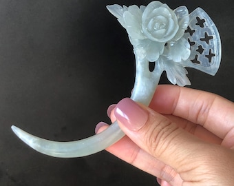 Puede incluir: Horquilla de jade verde pálido con diseño floral. El accesorio para el cabello presenta una rosa y hojas talladas, con un elemento en forma de abanico con detalles recortados. La varilla curva se sostiene en una mano.