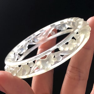 May include: A translucent white bangle bracelet featuring an intricate, carved floral pattern that wraps around the entire circumference. The material has a smooth, polished finish with subtle variations in opacity and light reflection throughout.