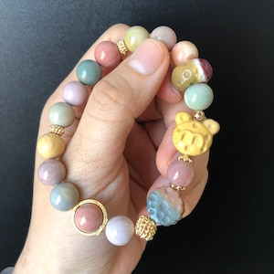 Könnte beinhalten: Ein mehrfarbiges Perlenarmband mit einem goldenen Anhänger in Form eines Bären. Die Perlen bestehen aus verschiedenen Steinsorten, darunter Achat, Jaspis und Quarz.