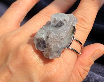 Può includere: Un anello d'argento con un grande cristallo di fluorite grezzo, grigio e viola, incastonato nella fascia.