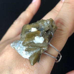 Può includere: Un anello realizzato con un cristallo grezzo, verde e bianco incastonato in una fascia d'argento.