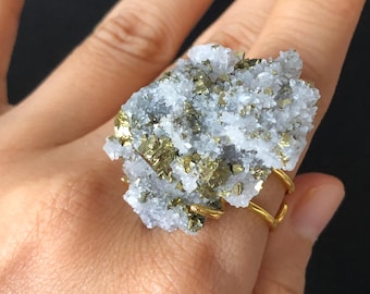 Könnte beinhalten: Ein Ring aus goldfarbenem Draht mit einem großen, natürlichen Cluster aus Pyrit- und Quarzkristallen. Der Ring wird an einem Finger gezeigt.