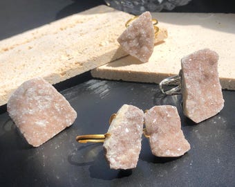 Puede incluir: Cuatro anillos con racimos de cristales rosados naturales engastados en bandas de metal dorado. Los anillos se muestran sobre una superficie marrón claro.