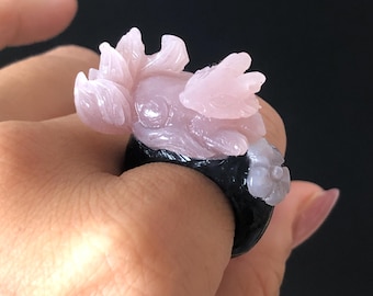 Puede incluir: Un anillo tallado con un diseño floral rosa claro sobre una banda negra. El diseño floral incluye pétalos y elementos en forma de alas. El anillo está hecho de piedra pulida.