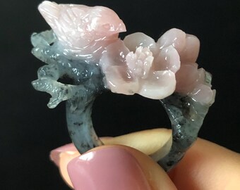 Puede incluir: Anillo de piedra tallada, con un pájaro posado en una rama con una flor. El pájaro y la flor son de color rosa suave, mientras que la rama y la banda del anillo son de color gris azulado moteado. El anillo es sostenido por una mano.