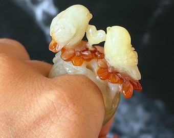 Puede incluir: Un anillo con dos pájaros posados en una rama con flores naranjas. El anillo está hecho de un material translúcido con detalles amarillos y naranjas. Los pájaros se miran.