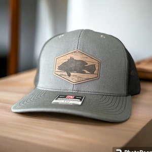 May include: Gray trucker hat with a black mesh back and a curved brim. The front panel features a wooden hexagon patch with a fish silhouette. The hat has a Richardson tag.