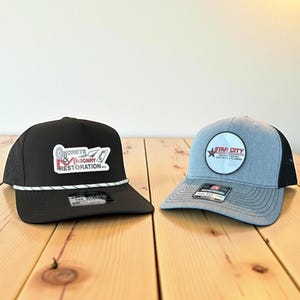 May include: Two baseball caps on a wooden surface. One is black with a white rope detail and a logo that reads "Concrete & Masonry Restoration." The other is gray with a black mesh back and a "Star City" logo.