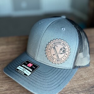 May include: A gray and black trucker hat with a brown leather patch that says "Measure Once Cuss Twice" with a saw blade and a tape measure graphic.
