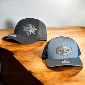 May include: Two black and gray mesh trucker hats with different logos. The first hat has a gray shield with the text "IRONCLAD" and "TOOL BELT CAUTION" inside. The second hat has a brown leather patch with the text "Your LOGO Here".