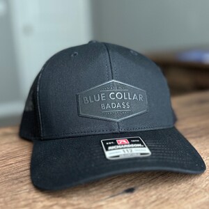 May include: Black trucker hat with a black leather patch that says "Made in America Blue Collar Badass". The hat is a Richardson 112.